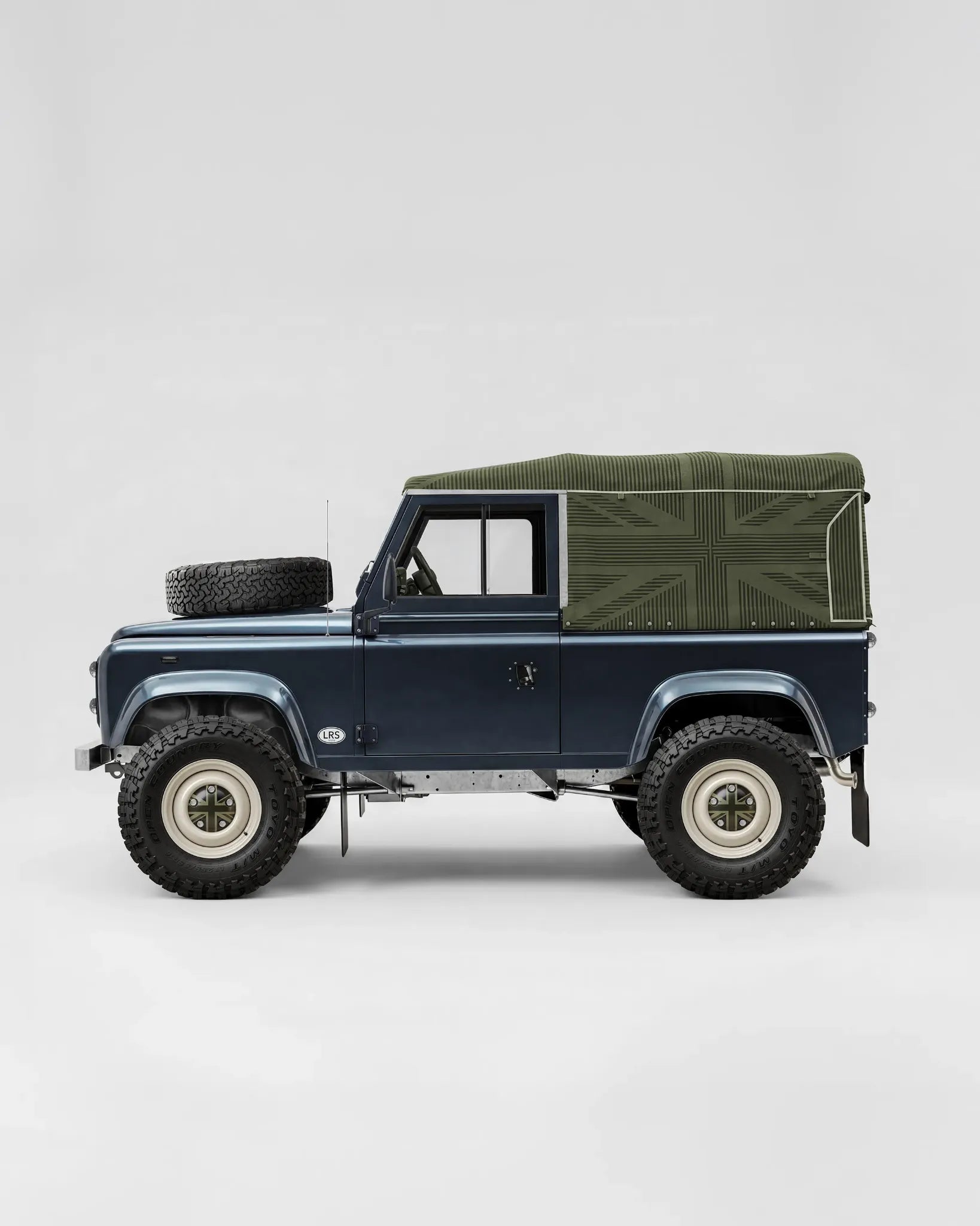 Side-profile design visualization of a blue Defender 90 with an olive Union Jack patterned canvas roof, light-colored wheels and bonnet spare wheel